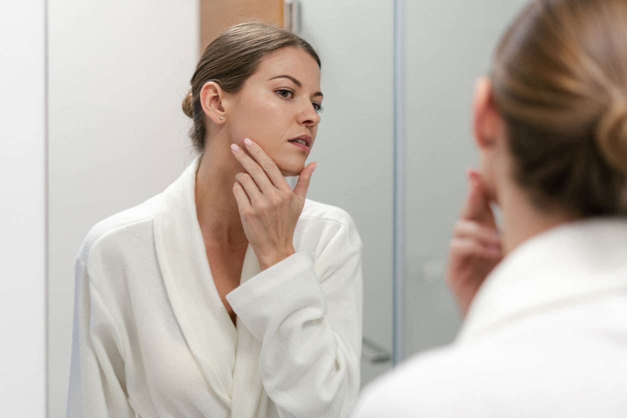 Stock image of a woman in bathrobe looking at her acne face in mirror